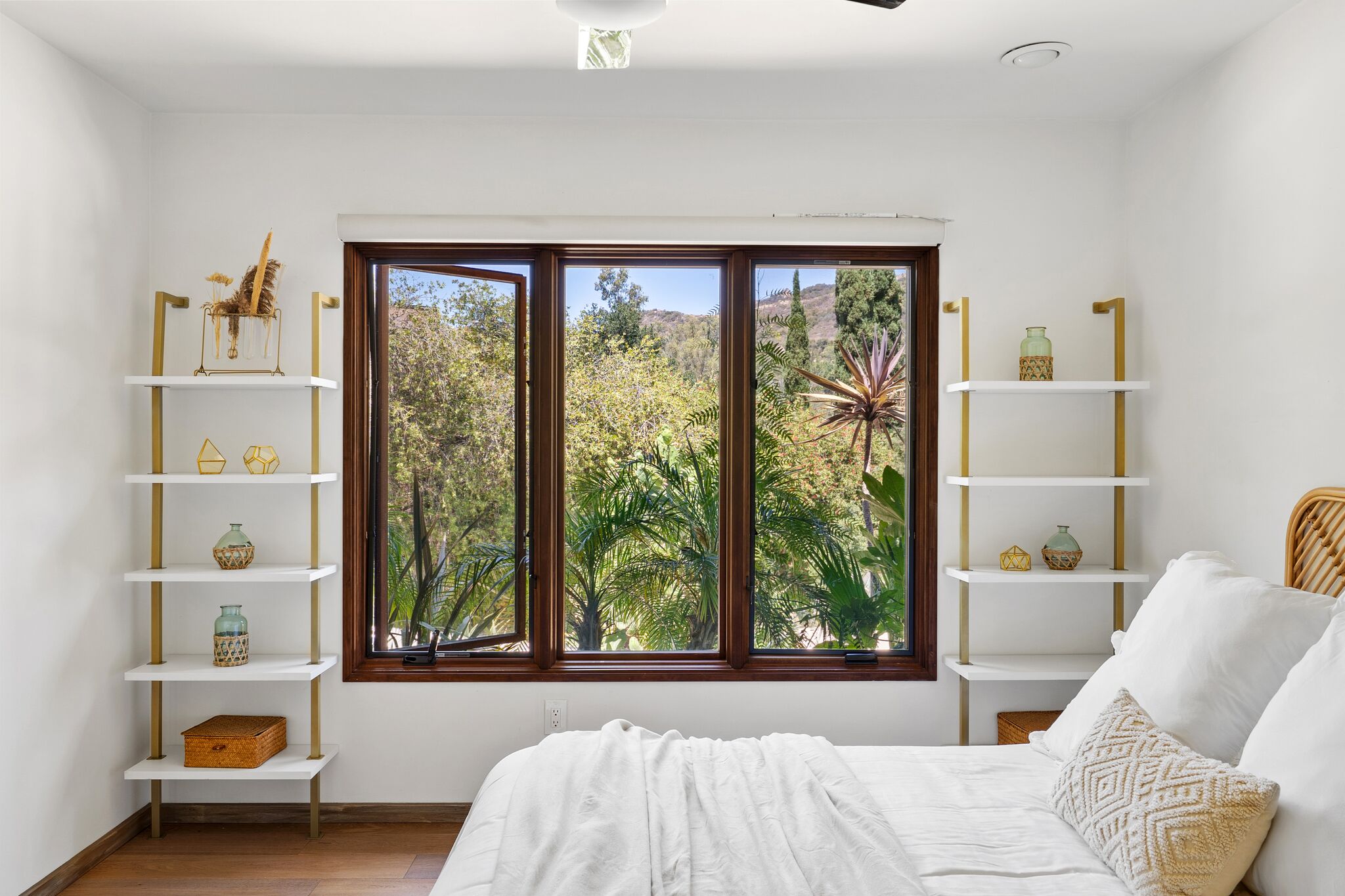 White bedroom with gold shelving and a wood-framed window full of palms and canyon hillside