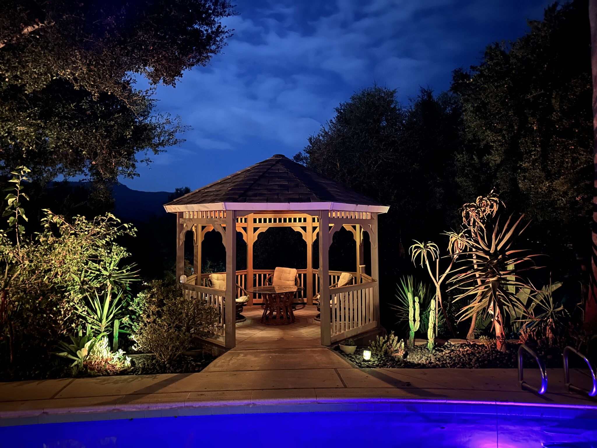 The gazebo glowing at night above the pool, lit cacti and palms against a deep blue sky
