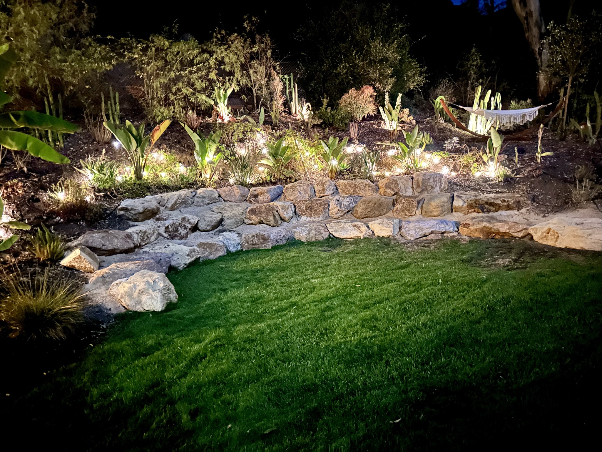 Boulder terrace glowing above the lawn at night with lit gardens and a hammock among the cacti
