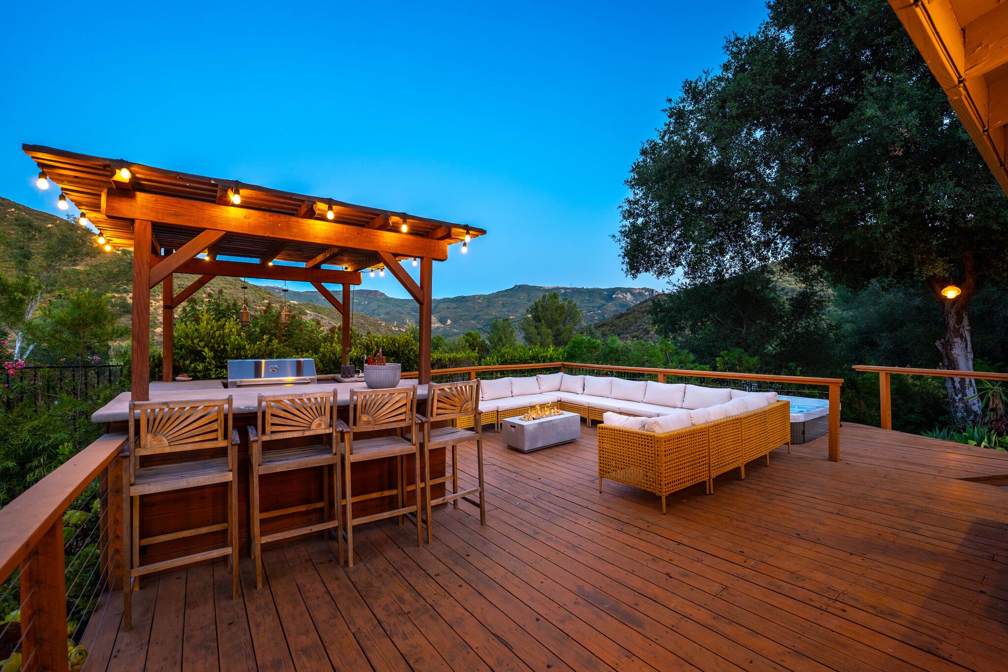 The full spa deck at blue hour: pergola bar with string lights, white sectional, and canyon ridgelines